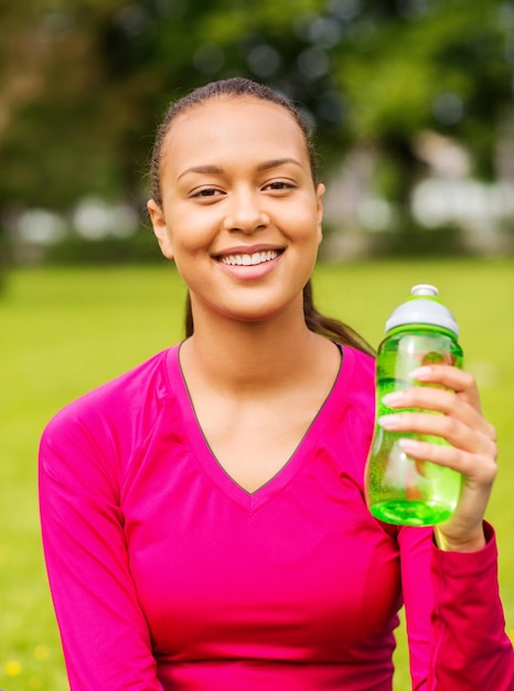 fitness-, park-, getränke- und sportkonzept - lächelnde afrikanisch-amerikanische frau, die draußen sitzt und flasche hält