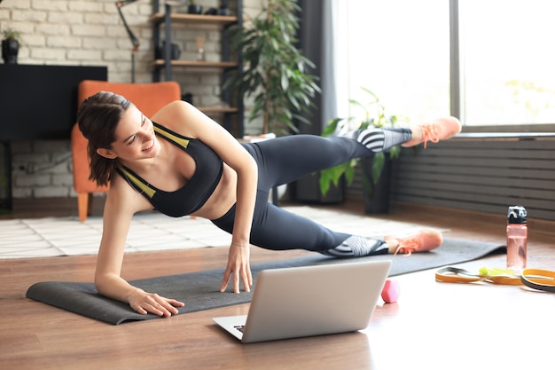 Fitness schöne schlanke Frau macht Side Plank und sieht sich Online-Tutorials auf dem Laptop an, trainiert im Wohnzimmer. Bleiben Sie zu Hause Aktivitäten.