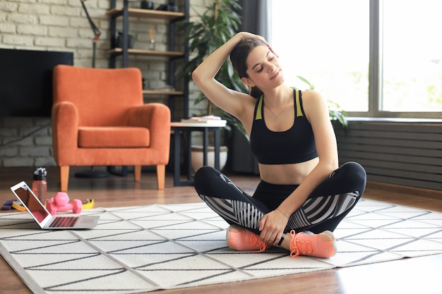 Fitness schöne schlanke Frau sitzt auf dem Boden mit Hanteln und einer Flasche Wasser mit Laptop zu Hause im Wohnzimmer. Bleiben Sie zu Hause Aktivitäten.