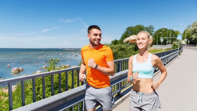 fitness, sport, freundschaft und gesundes lebensstilkonzept - lächelndes paar mit herzfrequenzuhr, die an der sommerküste läuft