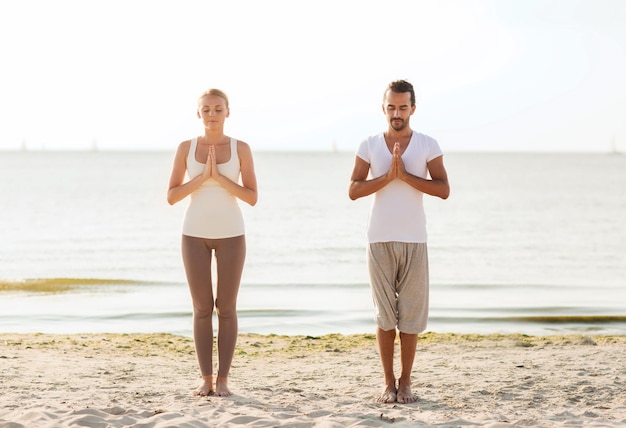 Fitness-, Sport-, Freundschafts- und Lifestyle-Konzept - lächelndes Paar, das Yoga-Übungen auf Sand im Freien macht