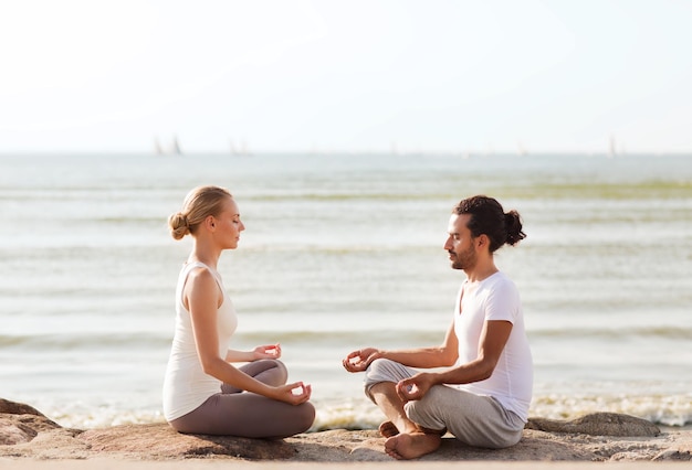 Fitness-, Sport-, Freundschafts- und Lifestyle-Konzept - lächelndes Paar, das Yoga-Übungen macht, die draußen auf Sand sitzen