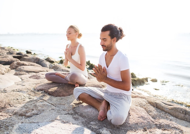 Fitness-, Sport-, Freundschafts- und Lifestyle-Konzept - lächelndes Paar, das Yoga-Übungen macht, die draußen auf Sand sitzen