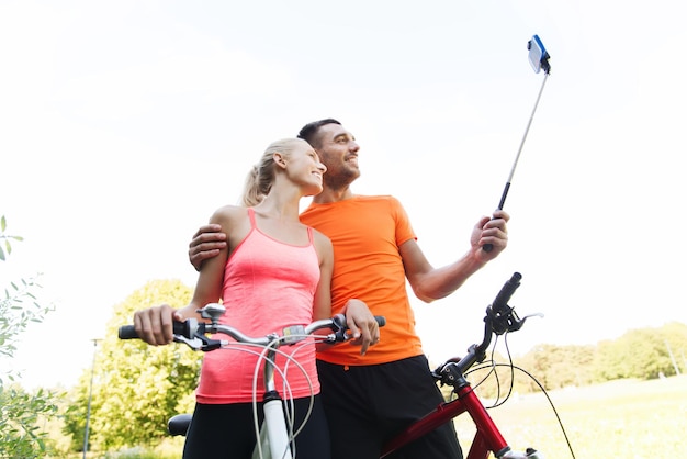 Fitness-, Sport-, Menschen-, Technologie- und gesundes Lebensstilkonzept - glückliches Paar mit dem Fahrrad, das mit dem Smartphone auf dem Selfie-Stick im Freien fotografiert