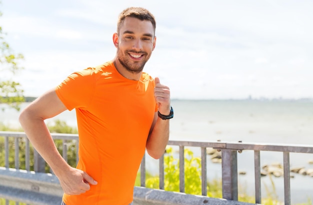 Fitness, Sport, Menschen, Technologie und gesundes Lifestyle-Konzept - lächelnder junger Mann mit Herzfrequenzuhr, der im Sommer am Meer läuft