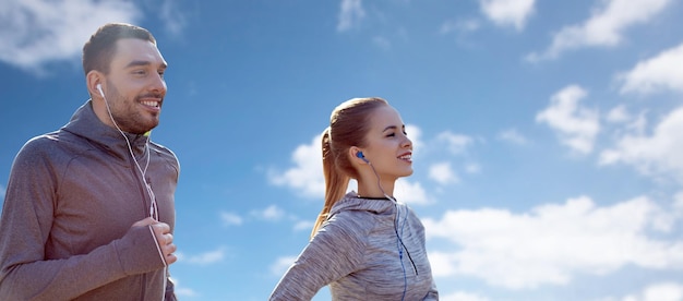 Fitness, Sport, Menschen, Technologie und Lifestyle-Konzept - glückliches Paar läuft und hört Musik in Kopfhörern über blauem Himmel und Wolkenhintergrund