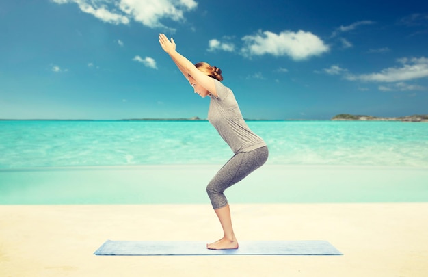 Fitness, Sport, Menschen und gesundes Lebensstilkonzept - Frau, die Yoga in Stuhlhaltung auf Matte über Strandhintergrund macht