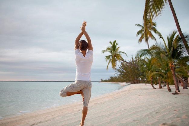 Fitness-, Sport-, People- und Lifestyle-Konzept - junger Mann, der von hinten Yoga-Übungen am Strand macht