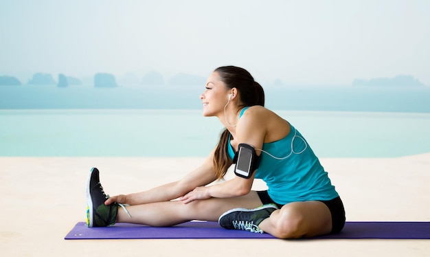 Fitness-, Sport-, Trainings-, Technologie- und People-Konzept - lächelnde Frau mit Smartphone und Kopfhörern, die Musik hört und das Bein auf der Trainingsmatte über Meer und Pool im Hintergrund des Hotelresorts streckt