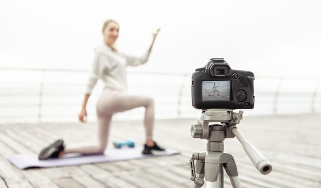 Fitnessbloggen. Junge gesunde Sportlerin zeichnet Yoga-Training vor der Kamera mit Stativ auf und zeigt, wie man Asana-Posen am Strand ausführt. Vloggen