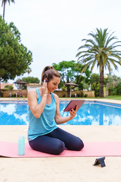 Fitnessfrau auf einem Online-Sportkurs im Freien. Vertikale Ansicht der jungen Frau, die sich mit Technologie zu einem Sportunterricht im Freien im tropischen Sommerrefugium neben einem Swimmingpool verbindet.