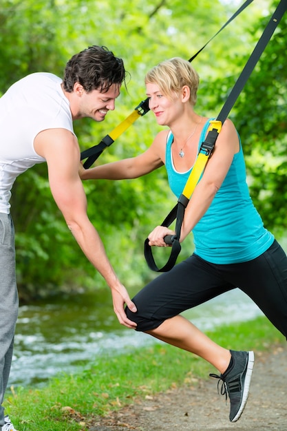 Fitnessfrau, die mit Suspensionstrainer und persönlichem Sporttrainer im Stadtpark unter Sommerbäumen trainiert