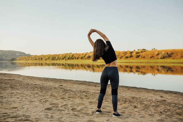 Fitnessfrauen, die sich an der Küste ausdehnen. Sportliche Frau mit perfektem schlanken Körper in der Modesportkleidung, die sich vor intensivem Training am Strand aufwärmt.