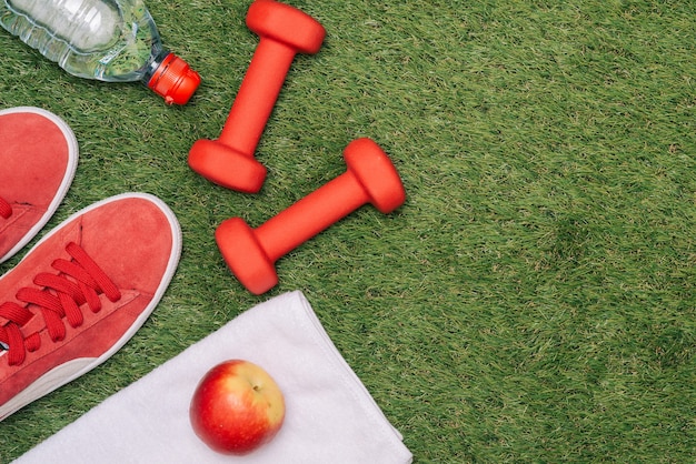 Fitnesskonzept mit Trainingsgeräten, Schuhen, Apfel, Hantel und Wasser auf grünem Gras.