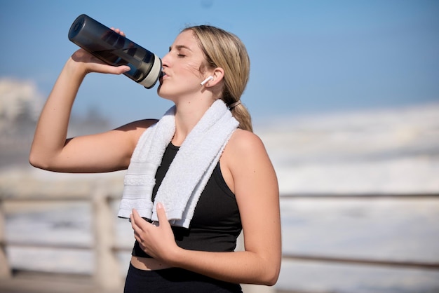 Fitnessmusik und eine Frau trinken Wasser am Strand für Sport, Laufen und Cardio-Routine. Podcast-Flasche und durstige Läuferin entspannen sich in der Trainingspause, während sie einen Online-Motivationstrack streamen