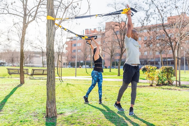 Fitnesspaar trainiert mit erhobenen Armen und dreht den Körper mit Trx-Fitnessgurten im Park. Multiethnische Menschen, die draußen trainieren.