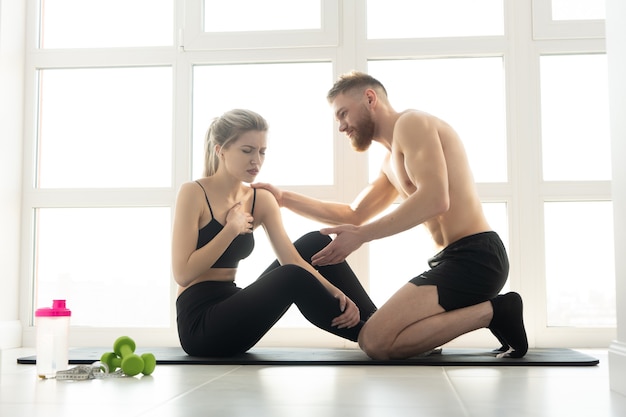 Fitnesstrainer und müde Sportlerin mit Herzschmerzen nach dem Training auf der Fitnessmatte. Blonde Frau trägt Sportkleidung. Mann mit nacktem Oberkörper. Konzept der sportlichen Aktivität zu Hause. Sonnige geräumige Wohnung