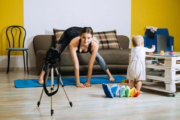 Fitnesstrainerin der jungen Mutter nimmt Video in der Nähe der Kamera zu Hause mit kleiner Tochter auf