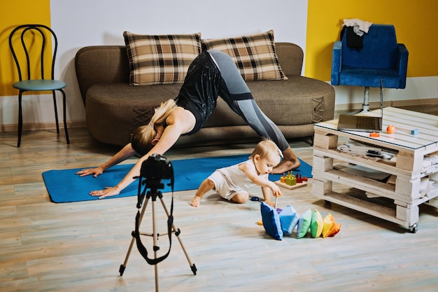 Fitnesstrainerin der jungen Mutter nimmt Video in der Nähe der Kamera zu Hause mit kleiner Tochter auf