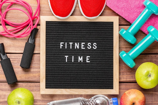 Fitnesszeit und Sportkonzept. Rote Turnschuhe, Äpfel, Springseil, Hanteln, rosa Handtuch und schwarzes Letterboard mit Worten Fitnesszeit auf Holzhintergrund.