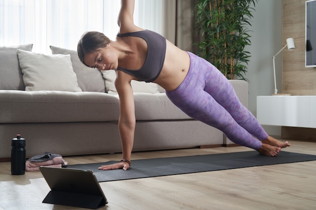 Fitte Frau, die Yoga auf ihrer Matte mit einem Online-Trainingsprogramm in ihrem Tablet praktiziert