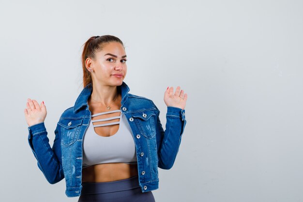 Fitte Frau in bauchfreiem Top, Jeansjacke, Leggings, die Handflächen in Kapitulationsgeste hebt und glücklich aussieht, Vorderansicht.