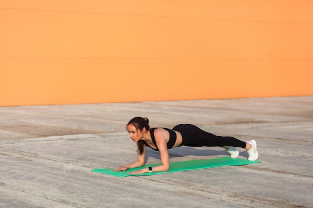 Fitte, schlanke, brünette Frau in engen Sporthosen, schwarzen Hosen und Top, die Yoga praktizieren, das in Ardha Phalakasana Low Plank Pose steht, trainiert Muskeln und Kraft, Sport im Gesundheitswesen im Freien