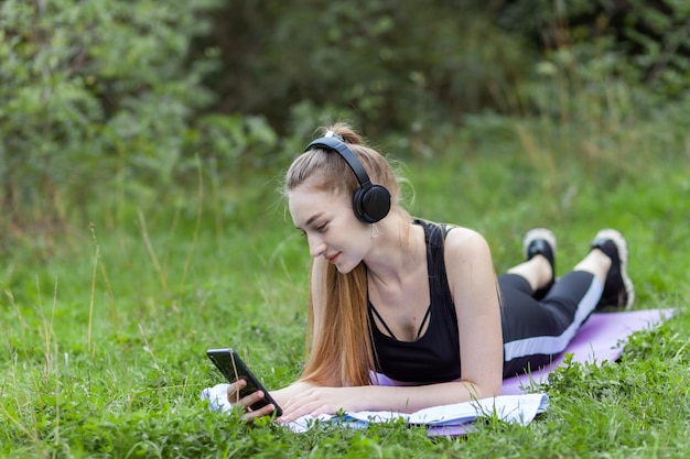 Fitte schlanke Frau liegt auf Matte, hört Musik über Kopfhörer und nutzt Smartphone im Park Gesunder Lebensstil