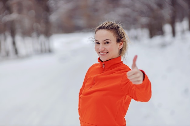 Fitte Sportlerin, die an einem verschneiten Tag in der Natur steht und Daumen nach oben zeigt. Genehmigen, gesunder Lebensstil, kaltes Wetter, Schnee, Winterfitness