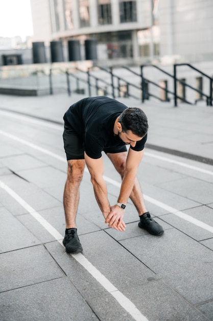 Fitter Mann, der sich vor dem Joggen zurückstreckt. Urbanes Sportkonzept. Männlicher Läufer dehnt sich aus und wärmt sich vor dem Joggen auf. Aktivitätskonzept am Morgen. Graue Stadt im Hintergrund.