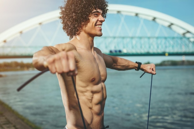 Fitter muskulöser junger Mann mit nacktem Oberkörper, der mit Widerstandsband in der Nähe des Flusses trainiert.