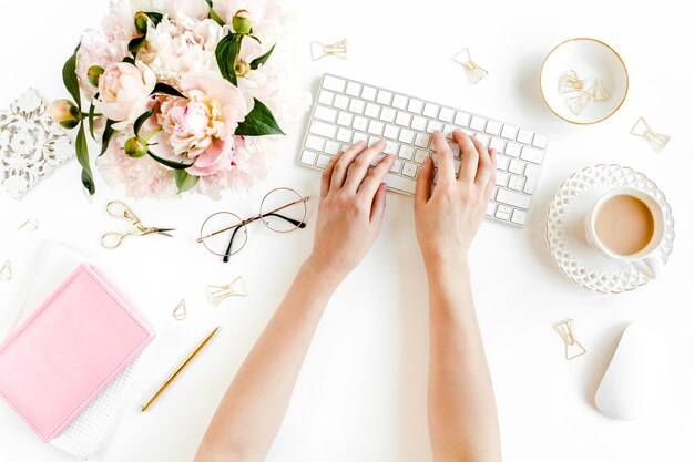 Flach gelegener Büro-Schreibtisch für Frauen Weiblicher Arbeitsbereich mit weiblichen Händen Computer rosa Pfeonen Bouquet Zubehör auf weißem Hintergrund Top-View