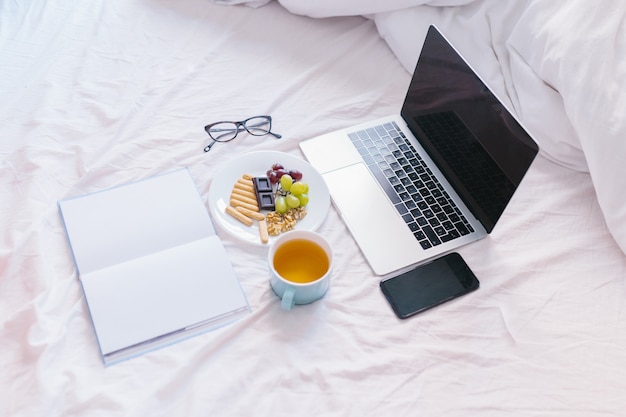 Foto flach liegen. tee zu hause in ihrem bett, ihren laptop überprüfen, ein buch lesen und frühstücken. draufsicht