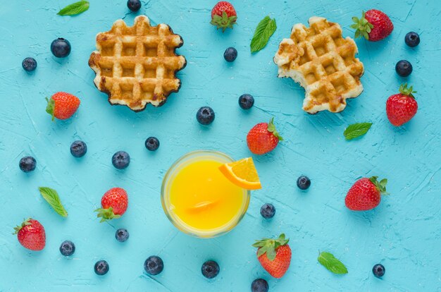 Flach liegen. Waffeln, Orangensaft, rote Beeren und Minzblätter auf blauer Oberfläche