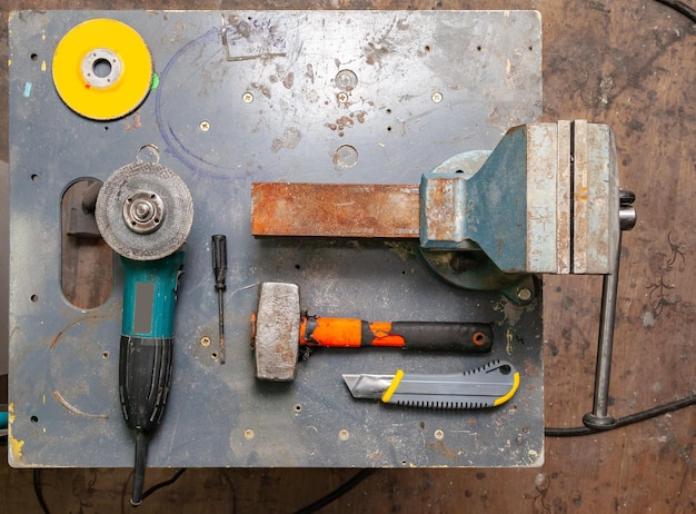 Flache Ansicht einer Werkbank mit einem Werkzeugsatz bestehend aus einem großen, schweren Schraubstock, Winkelschleifer, Schraubendreher, Schneider, Vorschlaghammer und einer Ersatztrennscheibe in einer Werkstatt