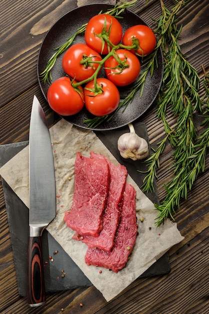 Flache Komposition mit rohem Rindfleisch, Küchenmesser, Tomaten und Rosmarin. Fleisch kochen.