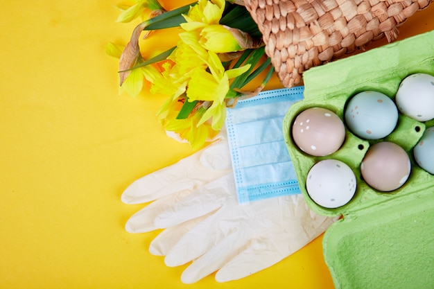 Flache Lage der Osterkomposition in der Coronavirus-Quarantäne. Ostersymbol Eier und Narzissenblumen im Korb nahe medizinischen Schutzmasken und Handschuhen