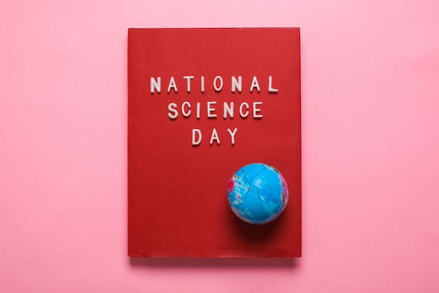 Flache Lage des National Science Day Schreibens auf rotem Buch mit Globus isoliert auf rosa Hintergrund