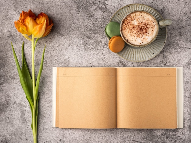 Flache Lage, Draufsicht auf weiblichen Schreibtisch. Kaffeetasse zum Frühstück, Makronen, leeres Notizbuch, orangefarbene Tulpe. Geschäftsplanung Frühstückskonzept.