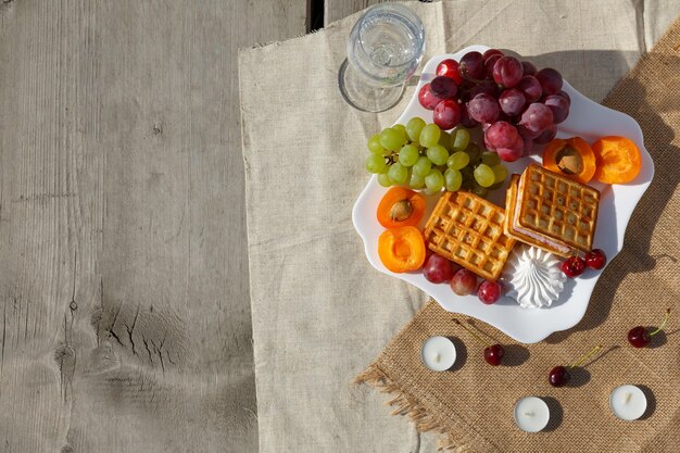 Flache Lage, Stillleben und Essensfoto. Picknick in der Natur an einem sonnigen Tag. Ein Gericht mit Früchten und Beeren, Waffeln und einem Glas sauberem Wasser steht auf einem Sackleinenstoff auf einem Holzboden aus alten Brettern