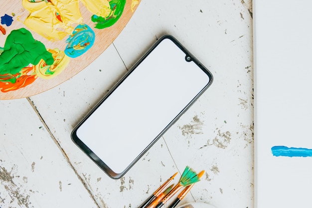 Flache Pinsel mit einem Smartphone-Leerraum und einer Farbpalette mit viel Pigment auf weißem Holzhintergrund. Hobby-Hintergrund. DIY, Basteldekoration für Herbstferien. Flache Lage, Ansicht von oben