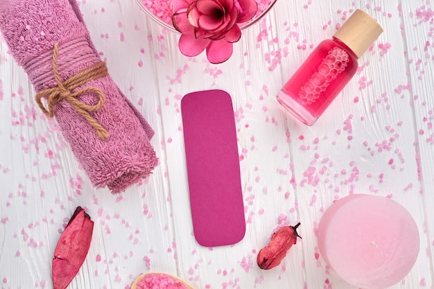 Flache rosa Spa-Accessoires auf weißem Holztisch. Gerolltes Handtuch mit Lotion, Kerze etc.