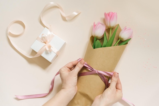 Flache weibliche Hände binden eine Satinbandschleife auf einem einfachen Strauß frischer rosafarbener Tulpenblumen in Bastelpapier