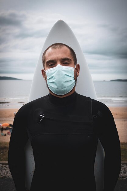 Flacher Fokus eines erwachsenen Mannes mit einer Gesichtsmaske und einem Surfbrett, das am Strand steht