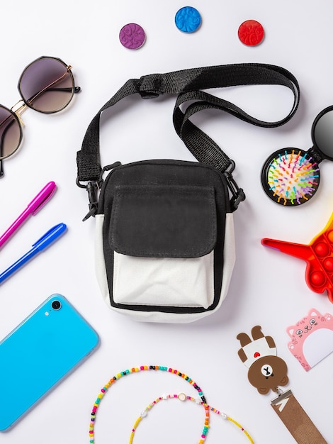 Flaches Foto der Babytasche mit Kosmetika und Smartphone-Pop-It-Spielzeug, Schlüsselanhänger, Sonnenbrille, Draufsicht auf grundlegende Schönheitsartikel für Mädchen