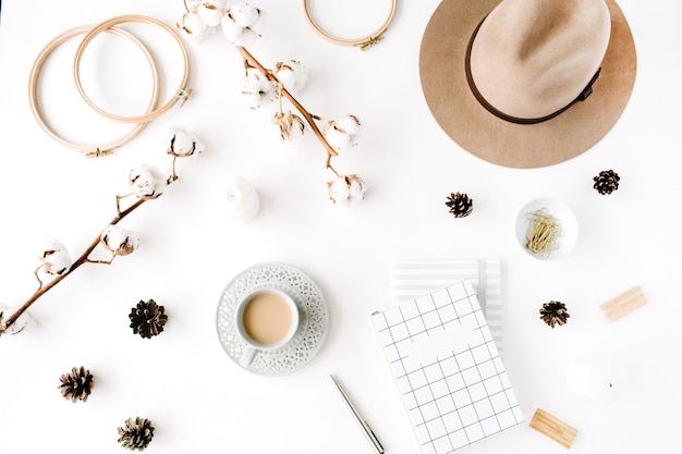 Flaches, trendiges, kreatives, feminines Accessoire-Arrangement mit Kaffee, Baumwollzweig und Tagebuch. Hut, Baumwollzweig, Notizbuch, Kaffeetasse, Tannenzapfen, goldene Clips auf Weiß