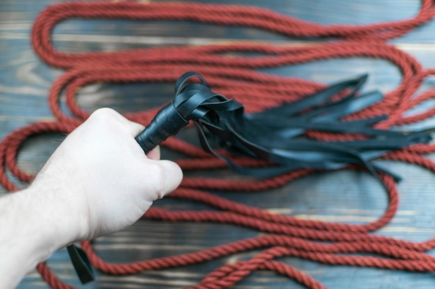 Foto flagelo para bdsm en la mano de un hombre y una cuerda para atar sobre un fondo oscuro accesorios para juegos sexuales