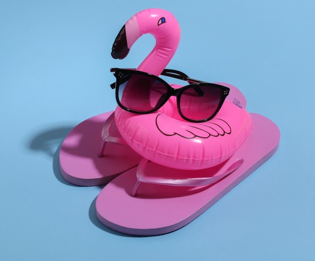 Flamenco rosado inflable con gafas de sol, chanclas sobre fondo azul soleado. Verano, concepto de vacaciones en la playa. Minimalismo.