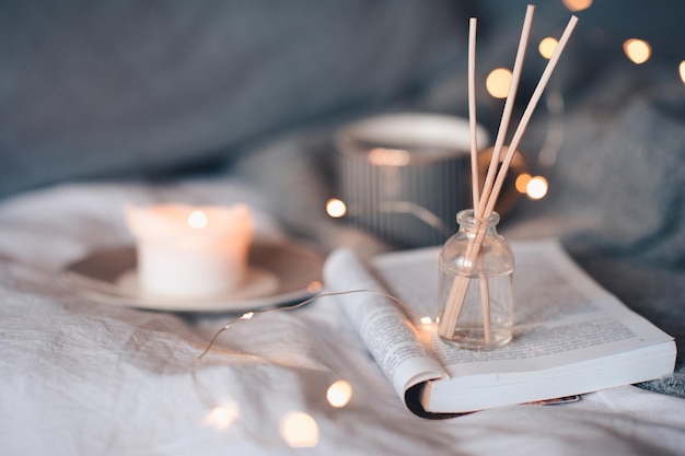 Flasche mit flüssigem Raumduft und hölzernen Bambusstäbchen, Düfte brennen Kerze und eine Tasse schwarzen Tee über Glühlichtern im Hintergrund aus nächster Nähe. Winterferienzeit. Gemütliche romantische Wohnatmosphäre.