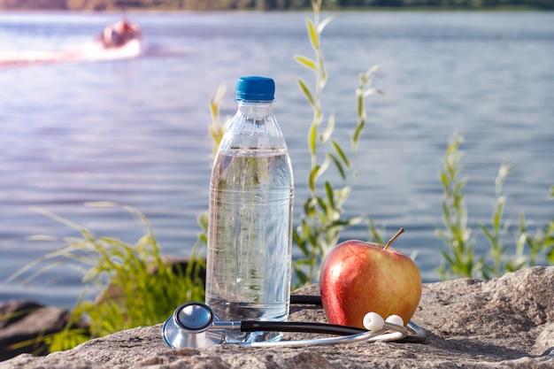 Flasche mit Wasser, Apfel und Stethoskop auf Felsen mit Flussdammhintergrund. Sommer, aktiver Lebensstil